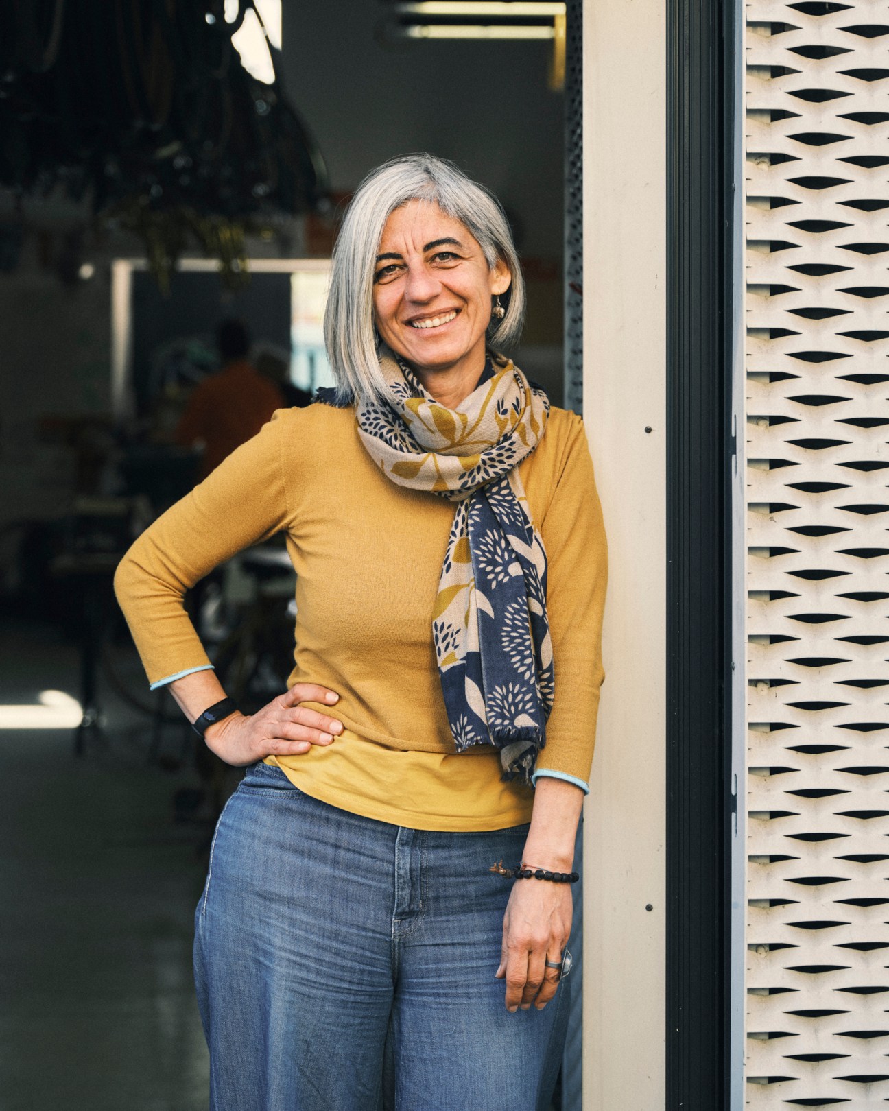 Silver-haired Micol Dell'Orto in yellow sweater and patterned scarf standing outside bike workshop