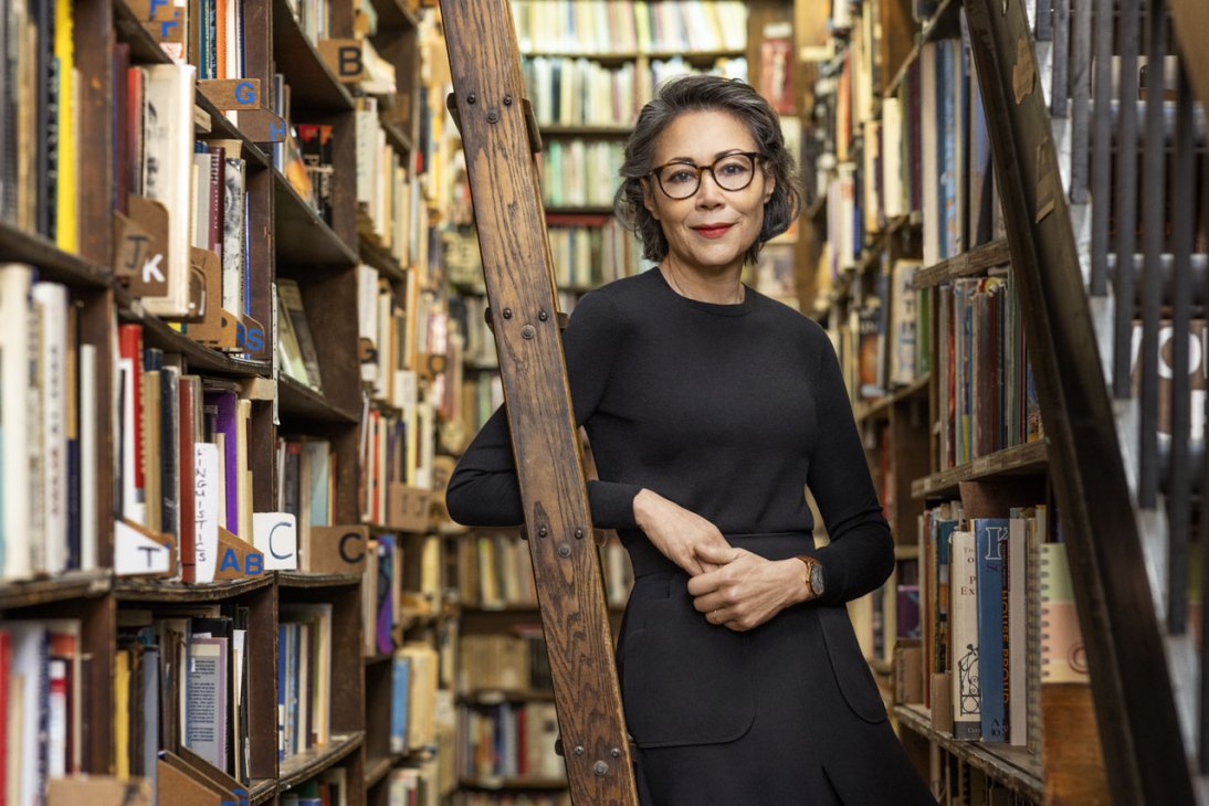 Ann Curry standing between tall bookshelves in a library wearing glasses and a black sweater