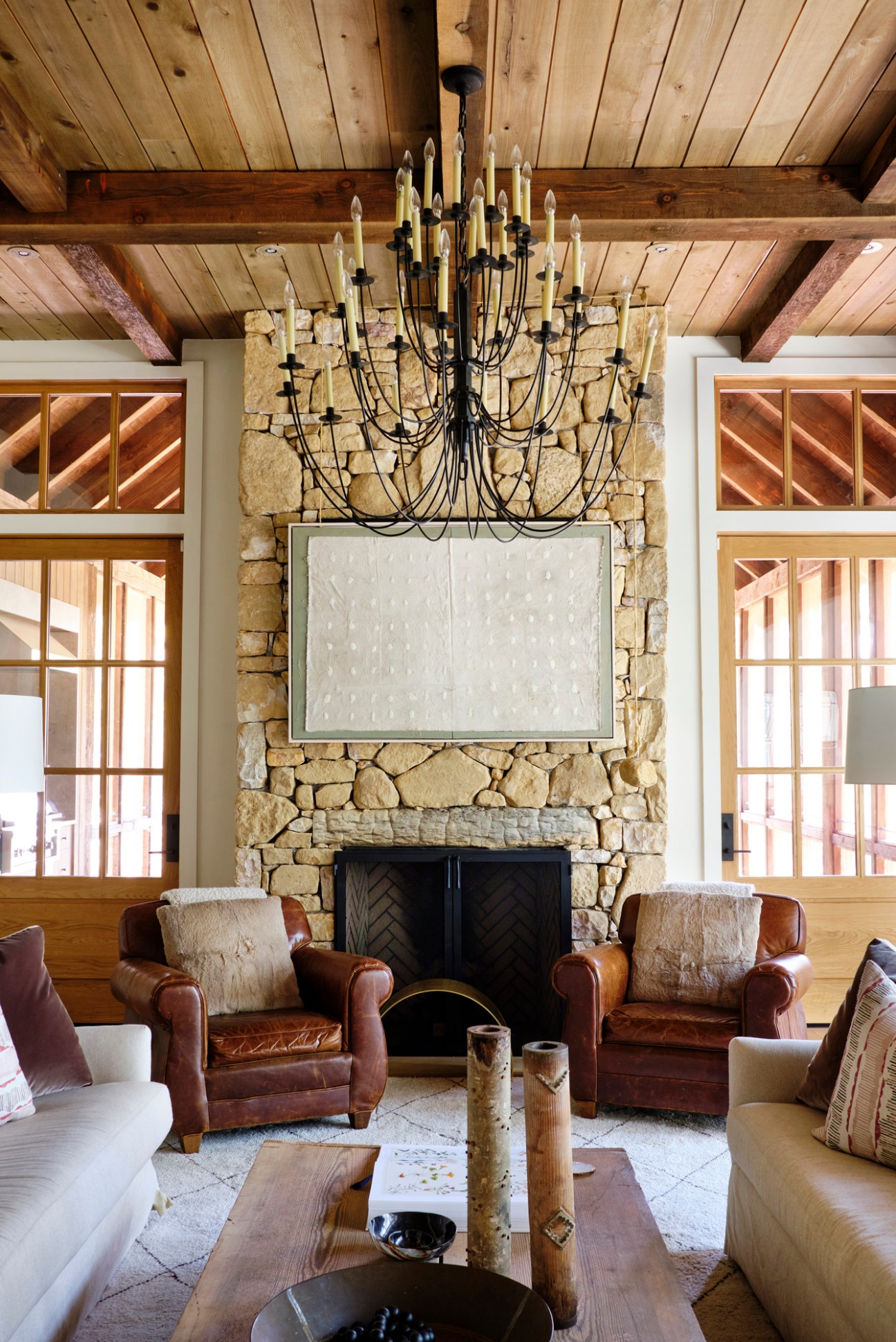 Stone fireplace with black wrought-iron chandelier in rustic lodge interior with wood beams and leather armchairs.