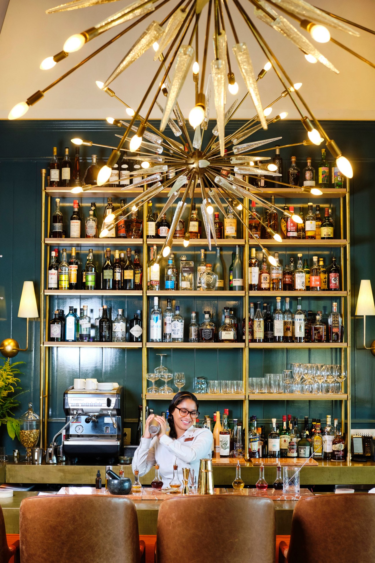 Bartender in white shirt shaking cocktail behind illuminated bar with stocked shelves of spirits and glassware.