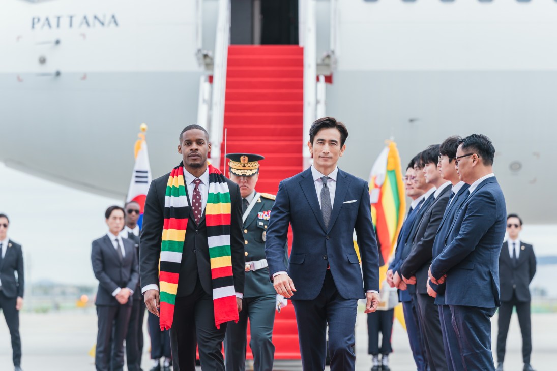 Two men in suits walking on red carpet at aircraft, one wearing colorful striped scarf