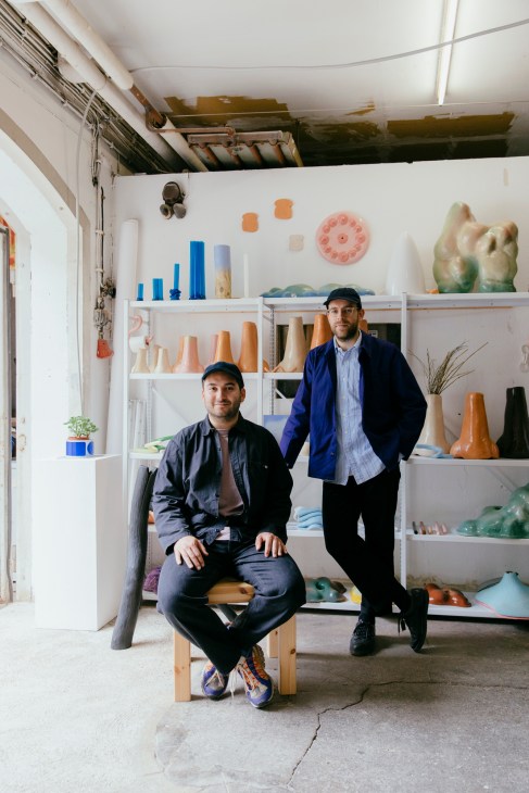 Two designers posing in their studio surrounded by shelves displaying ceramic vessels and design objects.