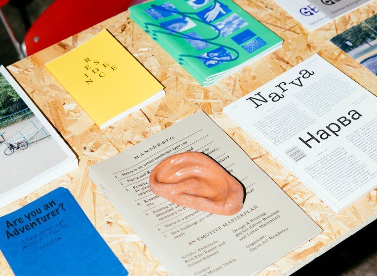 Design publications and objects displayed on OSB table with red stool, including yellow notebook, blue-green magazine, and terracotta sculptural piece.