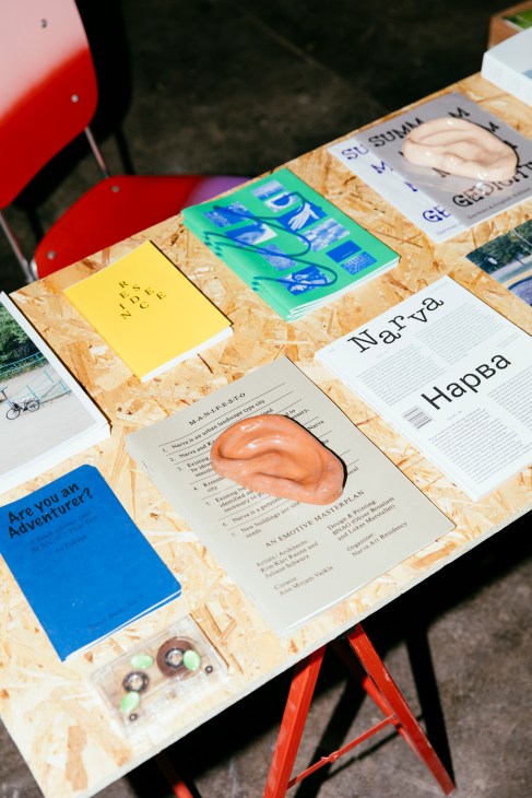 Design publications and objects displayed on OSB table with red stool, including yellow notebook, blue-green magazine, and terracotta sculptural piece.