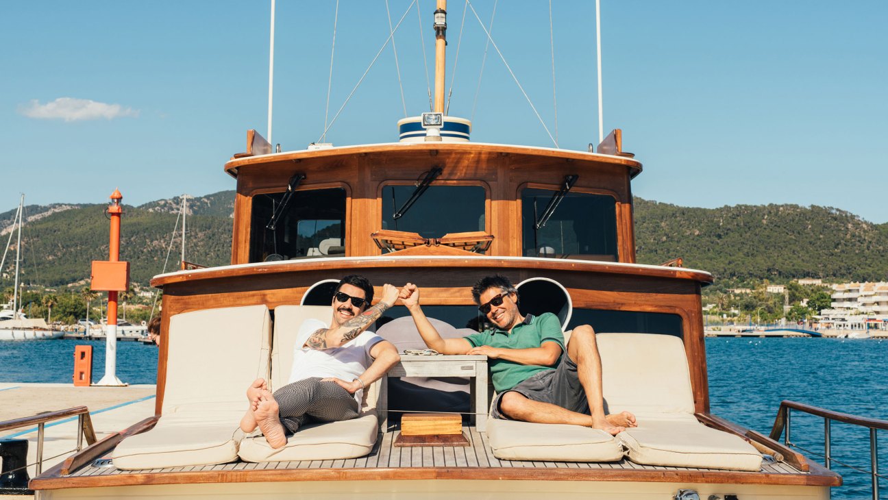 Two men relaxing on white cushioned seating aboard a wooden yacht with hills and harbor in background
