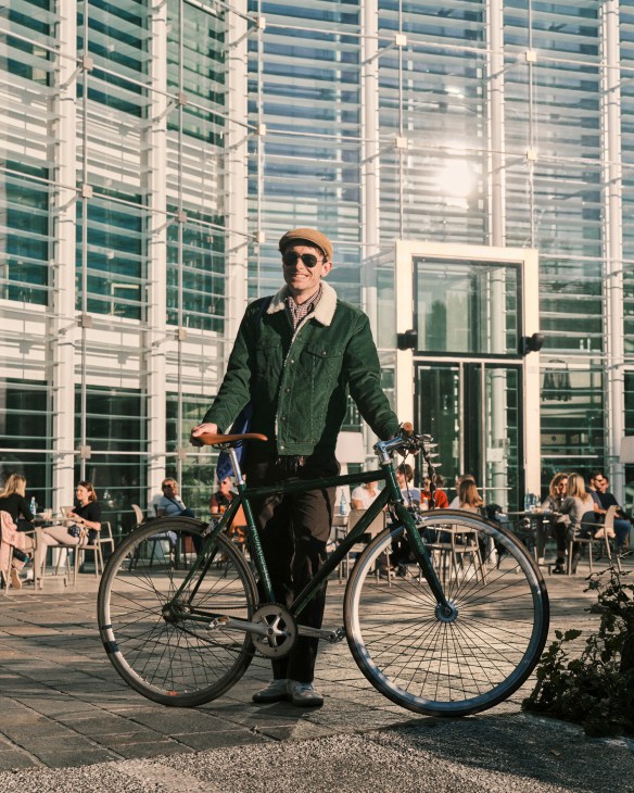 Man in green jacket and tan cap standing with bicycle in front of modern glass building