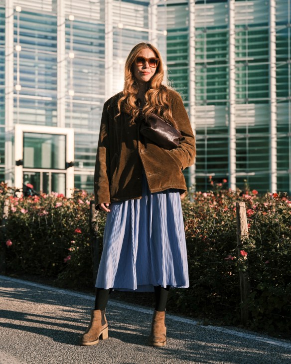 Woman in brown corduroy jacket and blue pleated skirt standing in rooftop garden with greenhouses behind