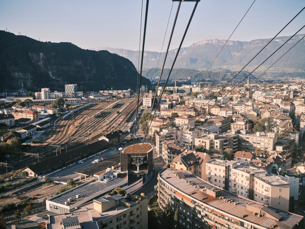 Elevated view of Bolzano with Dolomite mountains, railway yard, and pastel-colored buildings below cable car lines.