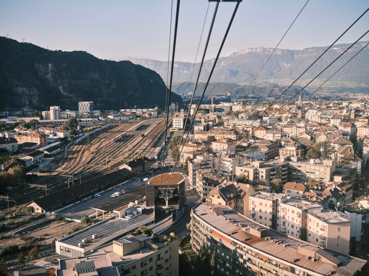 Elevated view of Bolzano with Dolomite mountains, railway yard, and pastel-colored buildings below cable car lines.