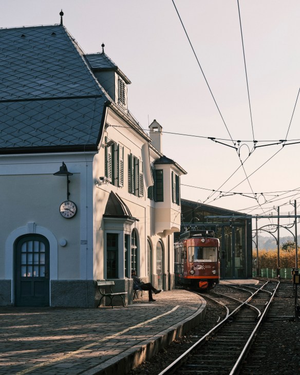 A red vintage tram passes historic buildings and railway tracks in Bolzano.