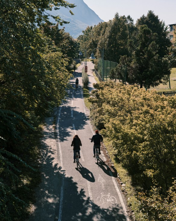 Two cyclists ride on a tree-lined path with Dolomite mountains visible in the background in Bolzano, Italy.