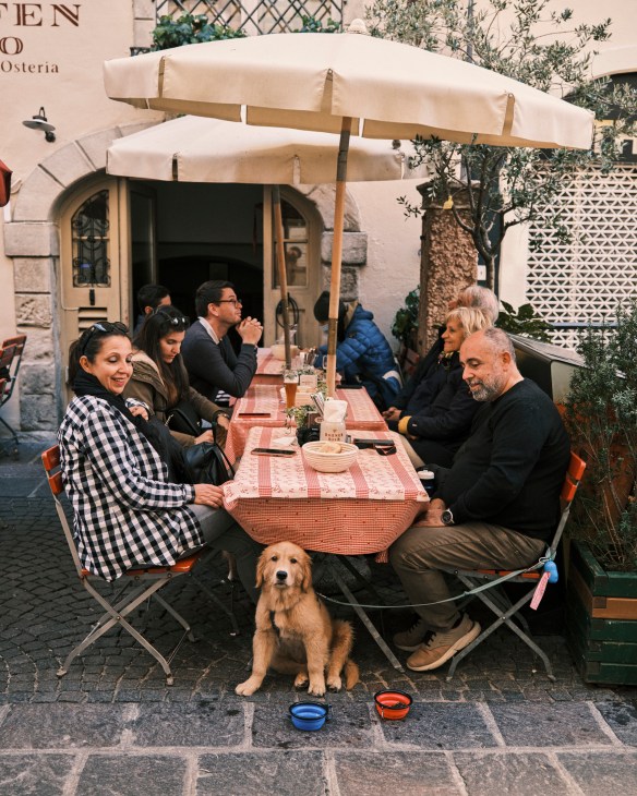 People dining at outdoor tables beneath cream umbrellas at a restaurant in Bolzano, Italy.