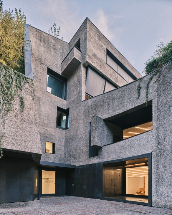 Modern concrete building with geometric windows and ivy vines, featuring illuminated ground floor openings.