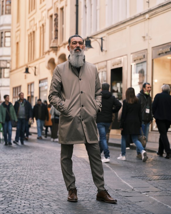 Man with long beard standing on cobblestone street in Bolzano with pastel buildings and pedestrians behind
