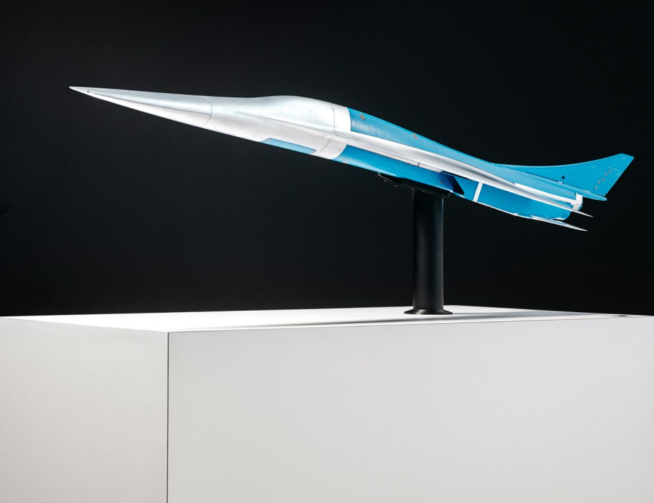 White and blue supersonic aircraft model displayed on black stand against dark background