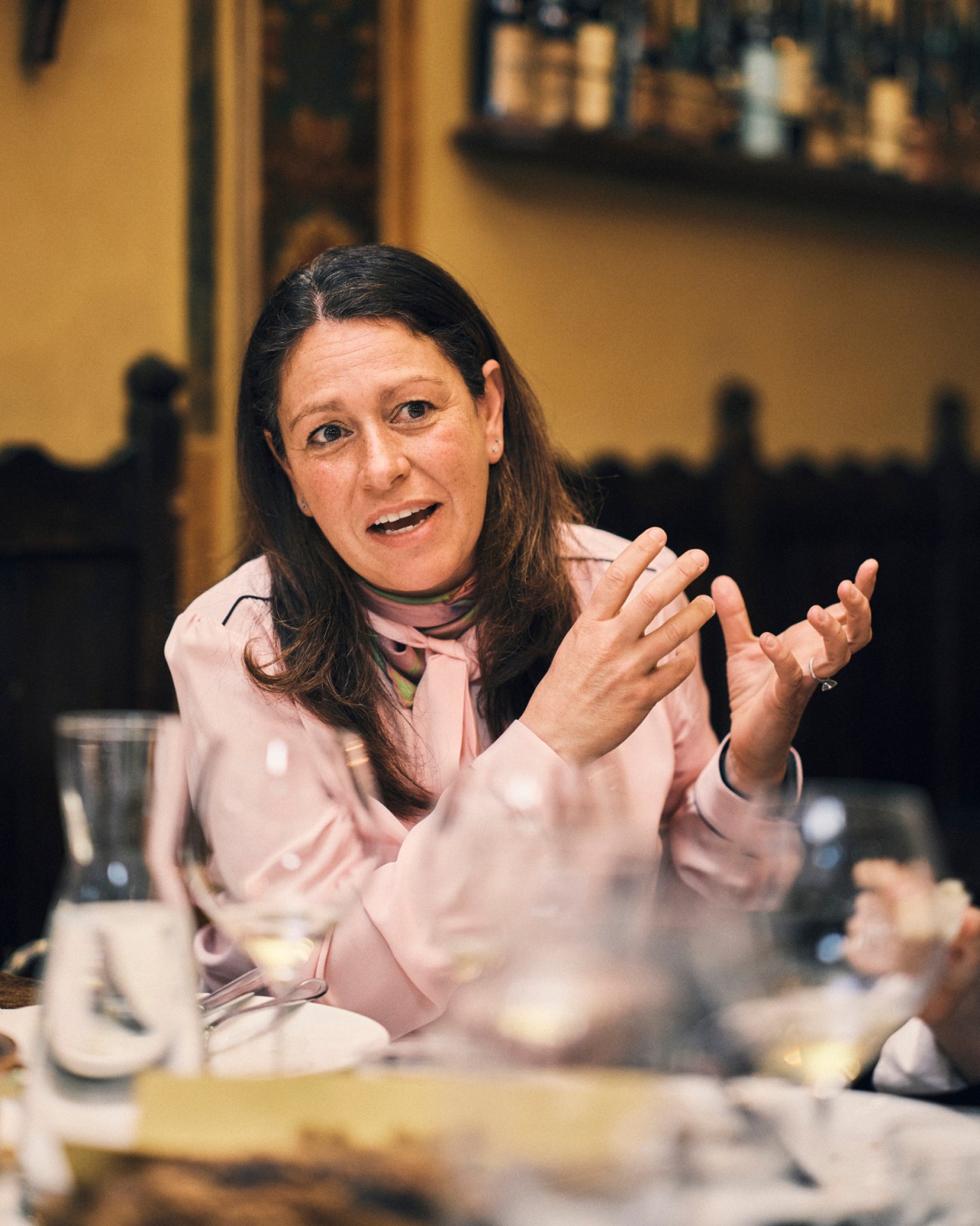 Woman with dark hair in pink jacket gesturing with hands while speaking at restaurant table