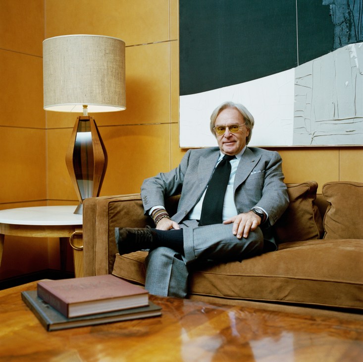 Man in grey suit and yellow glasses sitting on brown leather sofa in mid-century modern interior with yellow walls and table lamp.