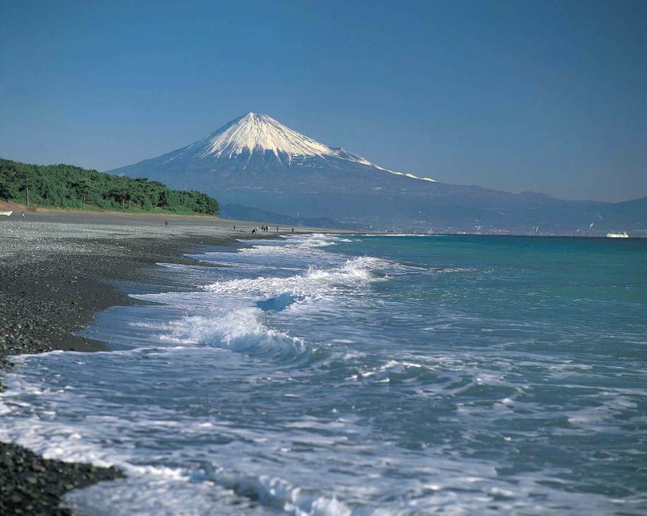 Mount Fuji snow-capped peak rises above a coastal beach with waves and forested shoreline.