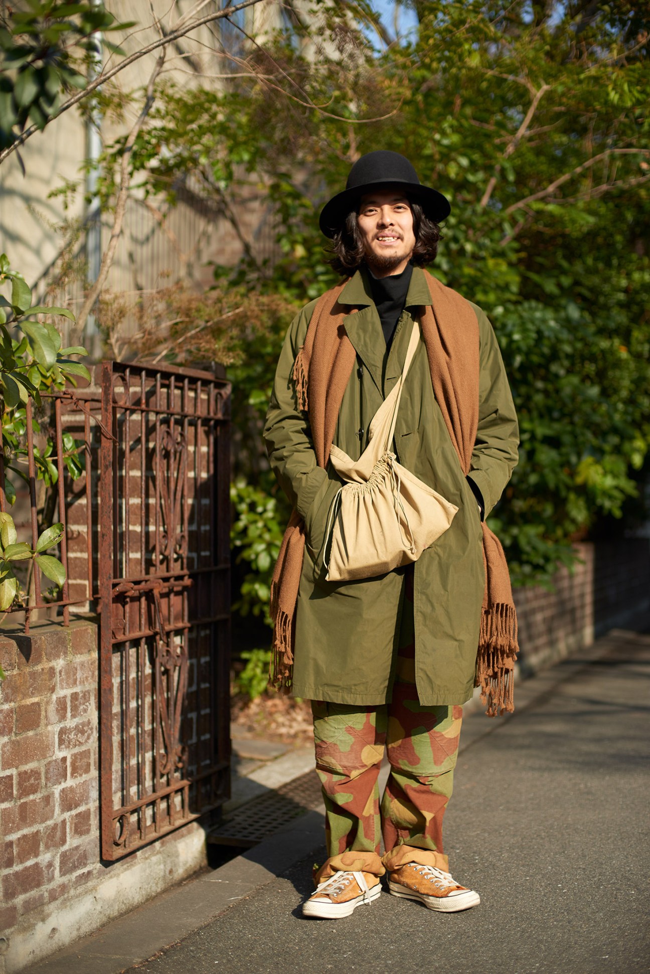 Man wearing olive coat, black hat, and patterned pants stands on a sunny street lined with ivy-covered buildings and a decorative gate.