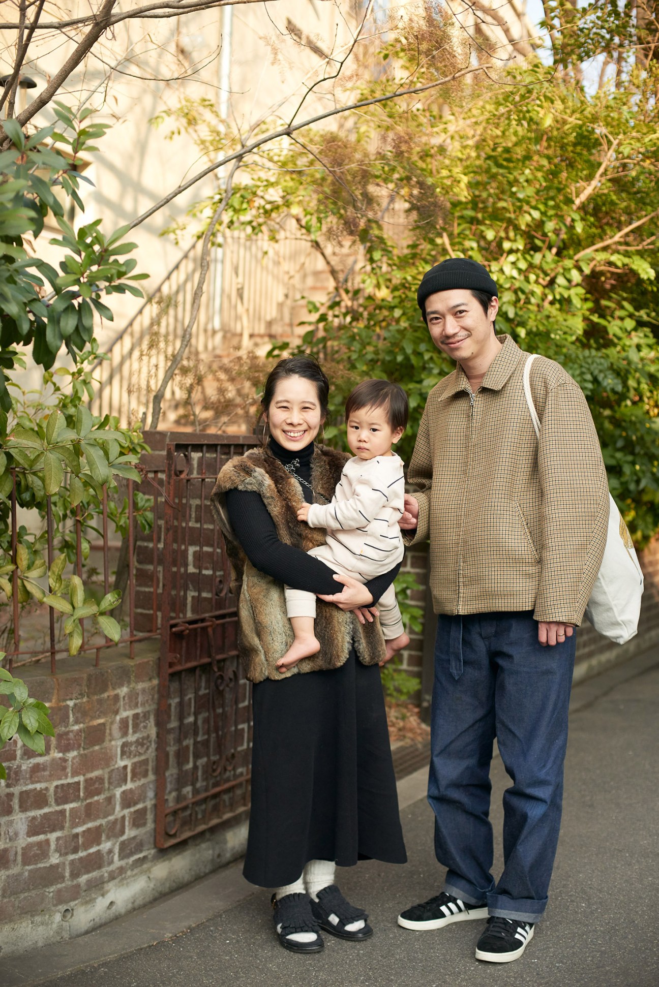 A young family of three standing in an alley with ivy-covered walls and a metal gate behind them.