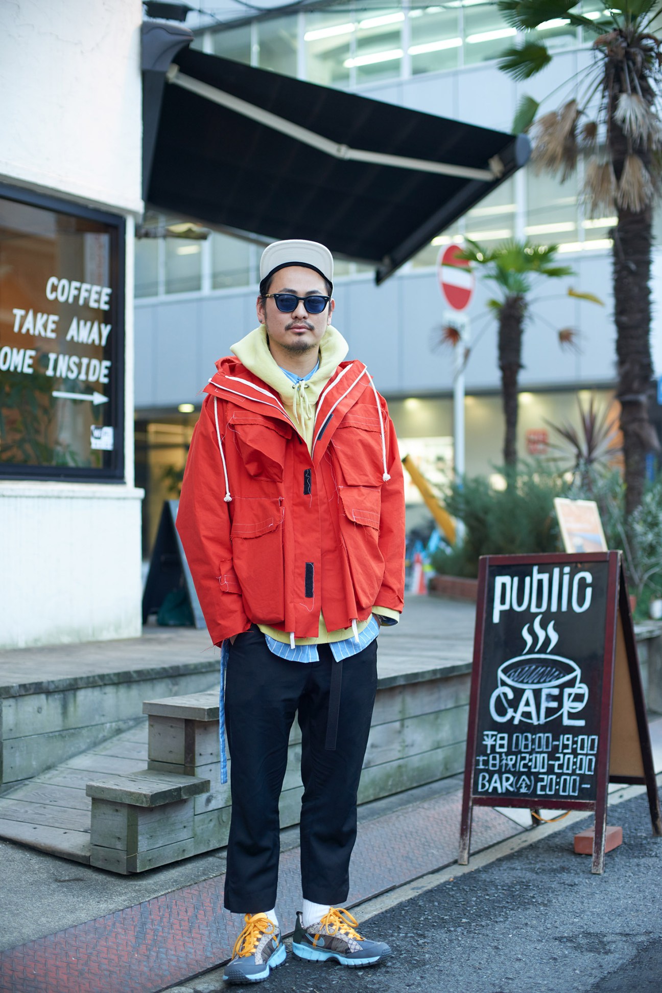 Man wearing red jacket and white cap stands outside a cafe with palm trees visible in background.