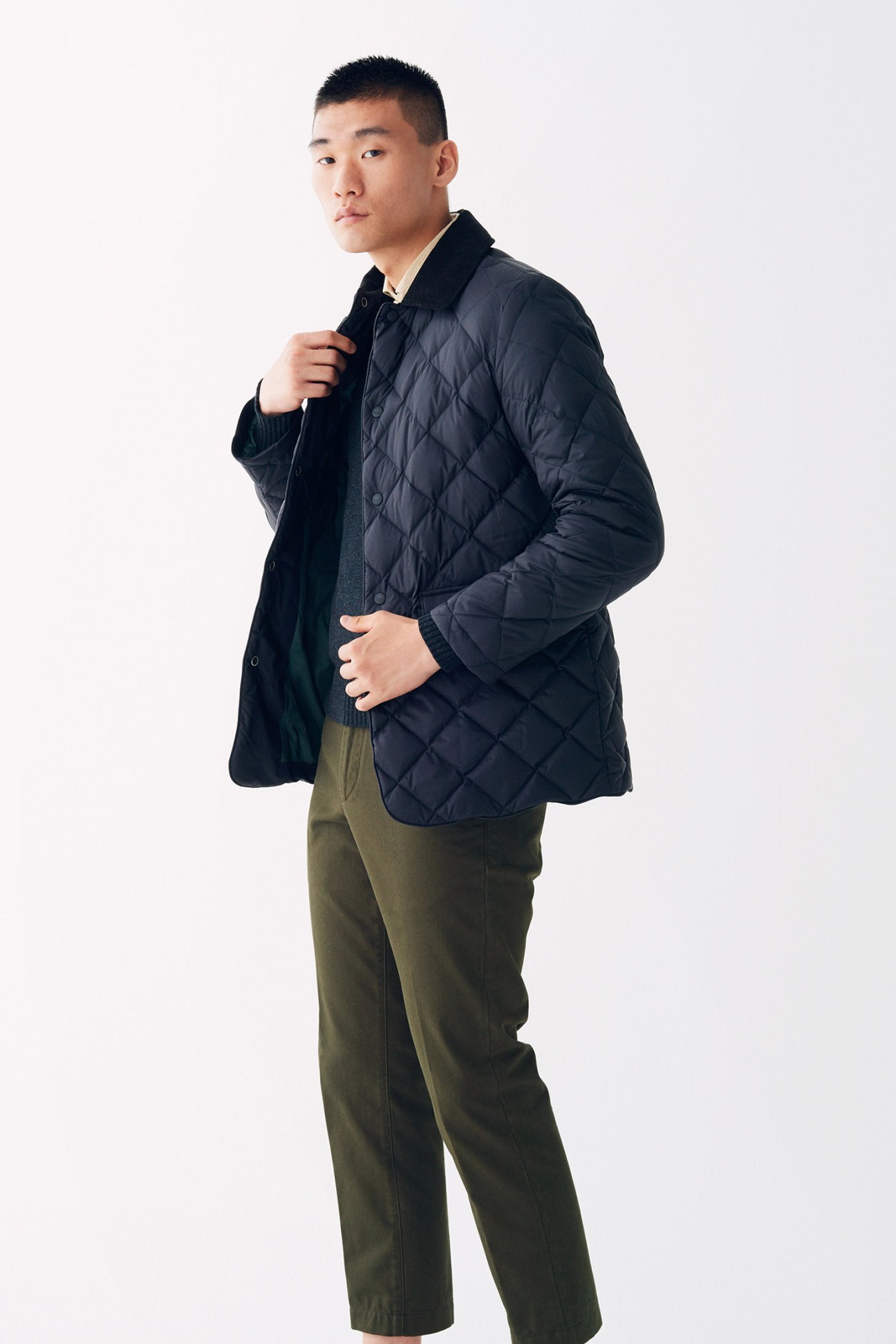 Man wearing navy quilted jacket with black collar over olive chinos against white background.