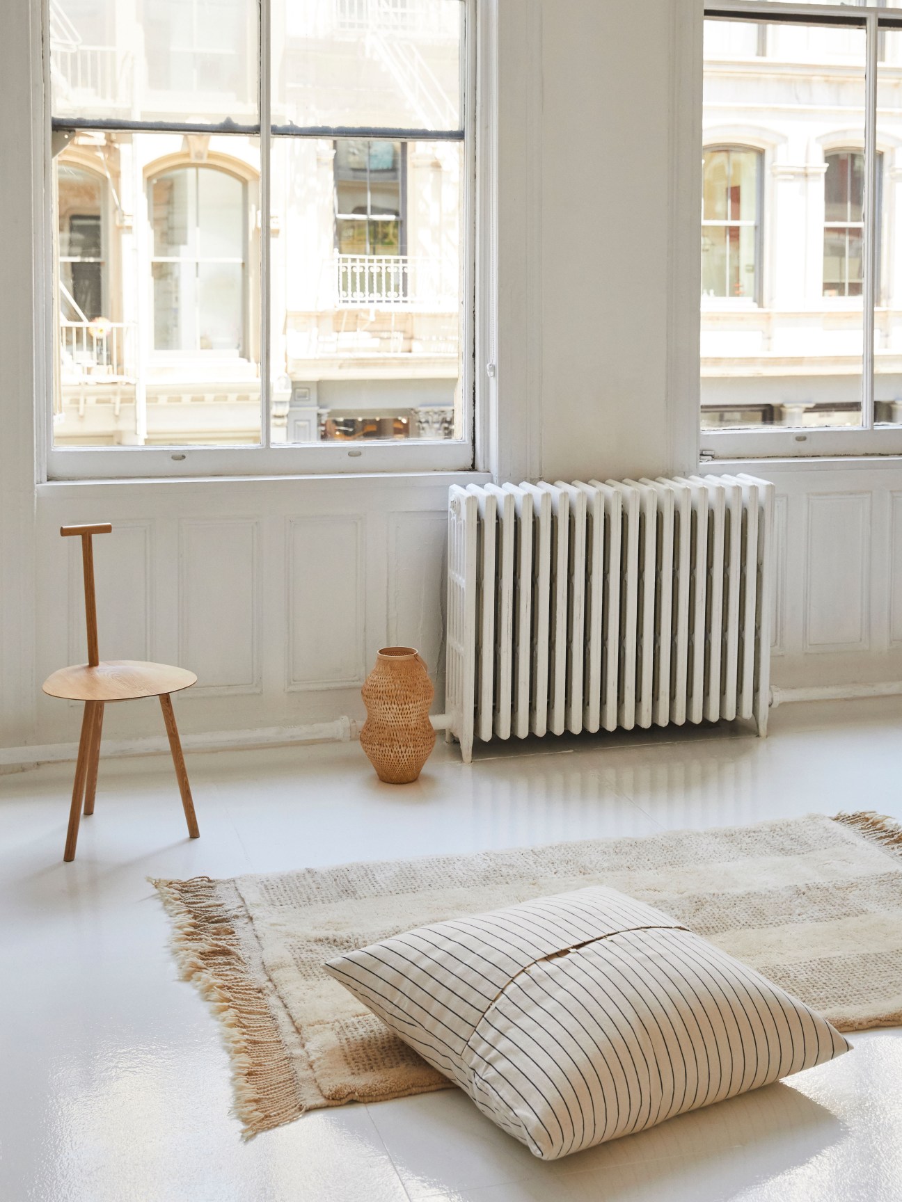 Minimalist interior with wooden chair, woven basket, striped pillows on rug, and white radiator by windows.