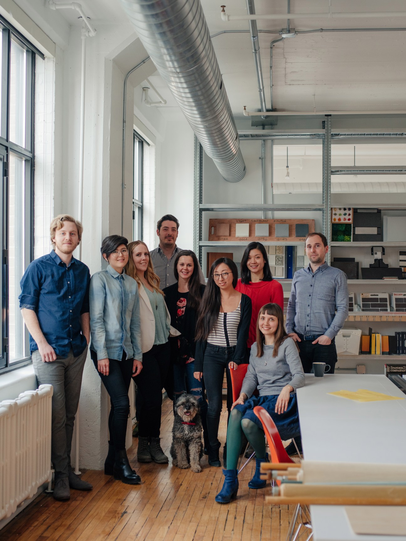 A group of nine people and a dog pose together in a bright studio space with industrial shelving and exposed ductwork.