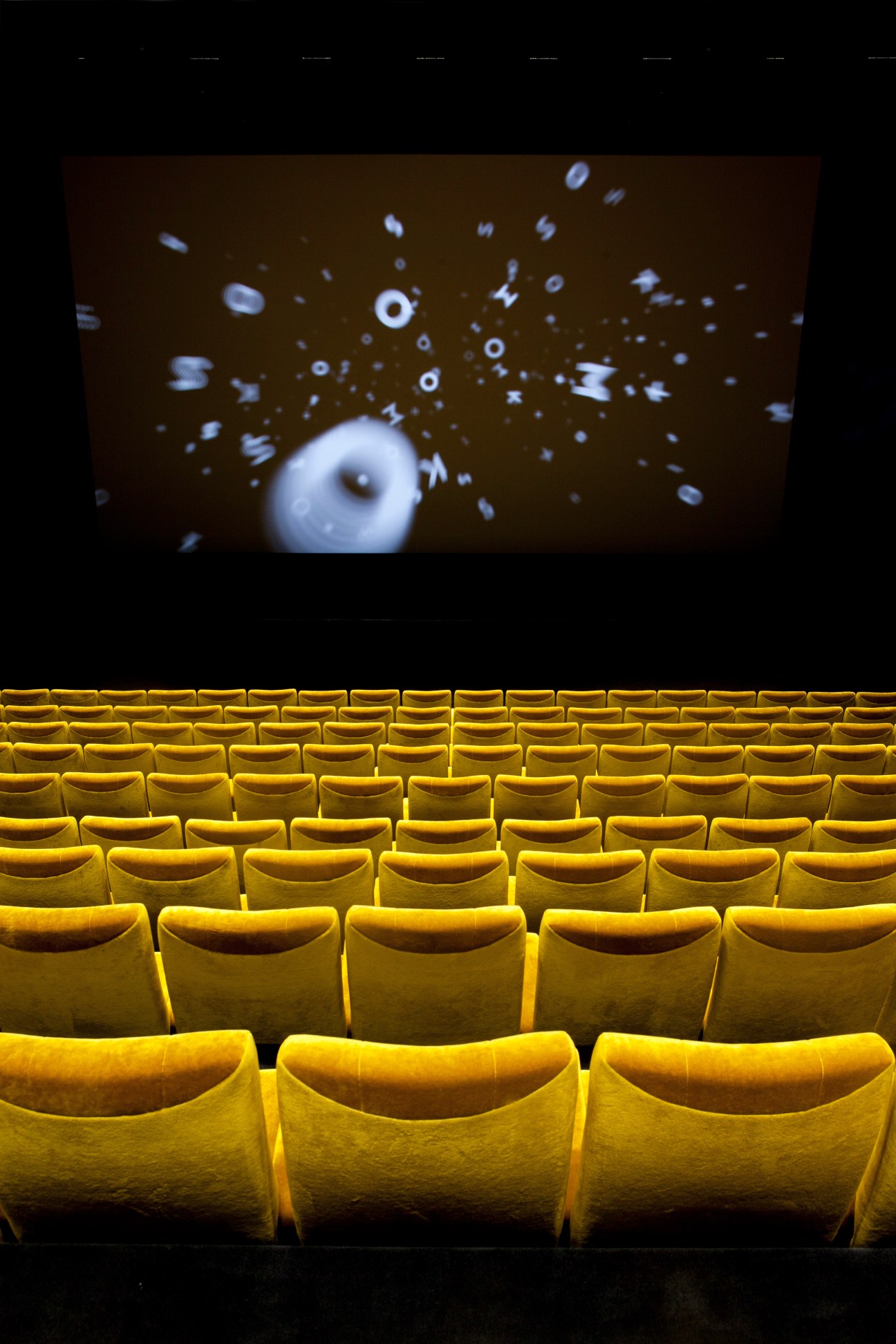 Yellow cinema seats facing a screen displaying an abstract white spherical object with scattered particles.