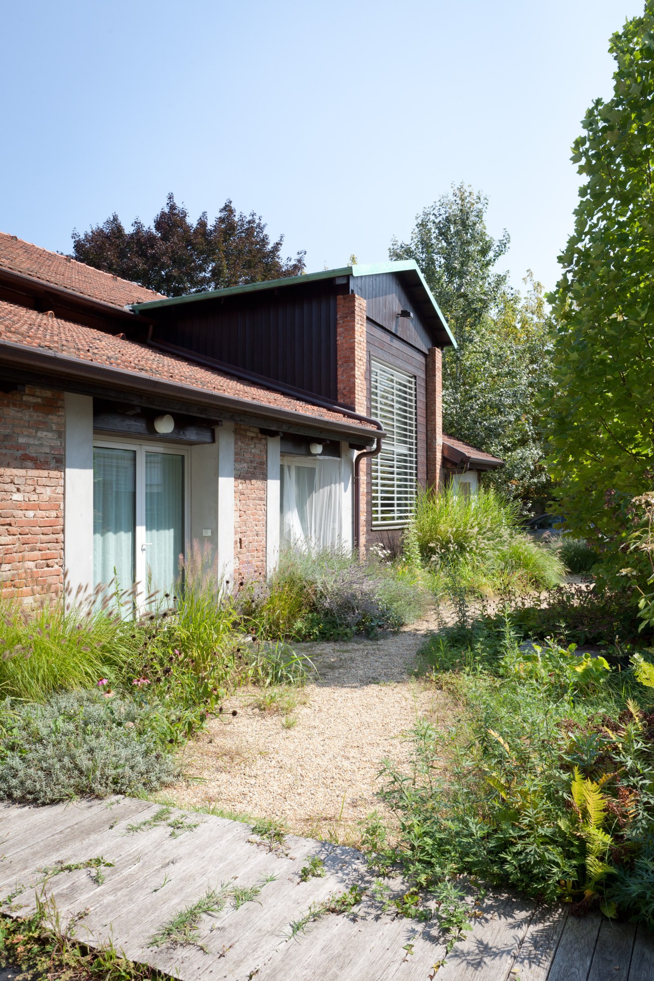 Modern brick house with black metal cladding addition and large windows surrounded by mature landscaping and gravel pathway.