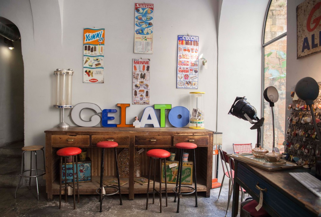 Vintage shop interior with colorful GELATO sign on wooden counter, red bar stools, and vintage posters on white walls.
