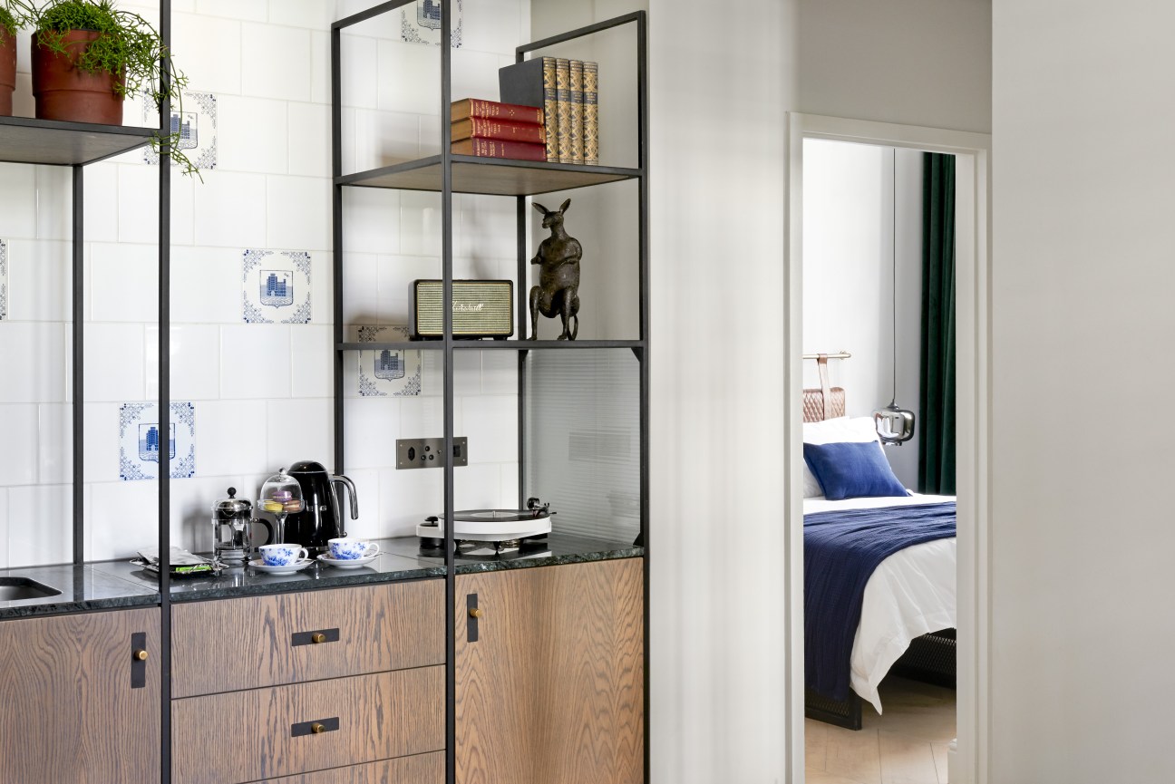 Hotel room kitchenette with wood cabinetry, black metal shelving, and view through to bedroom with blue bedding.