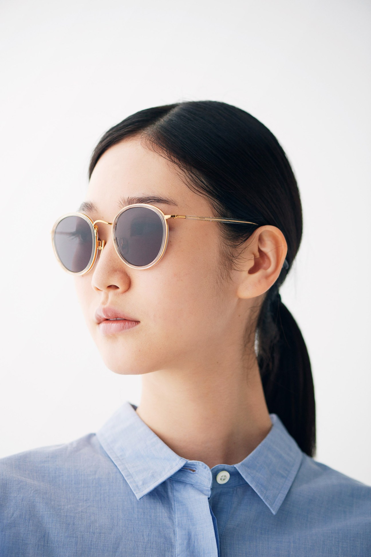 Woman wearing round-frame sunglasses with gold temples and gray lenses in a blue shirt.