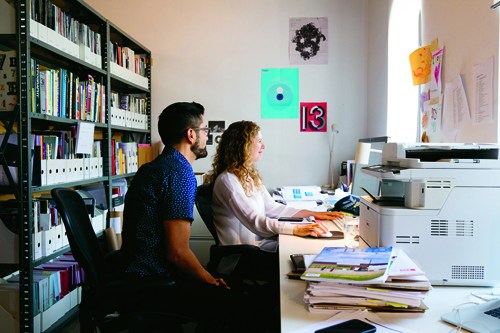 Two people working at desks in a bright office space with bookshelves and a printer visible.