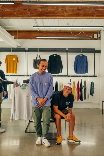 Two men pose inside a retail clothing store with shelves of folded garments and hanging clothes visible behind them.