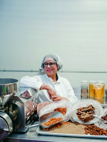 Woman wearing hairnet and white coat with spice containers and processing equipment in background.
