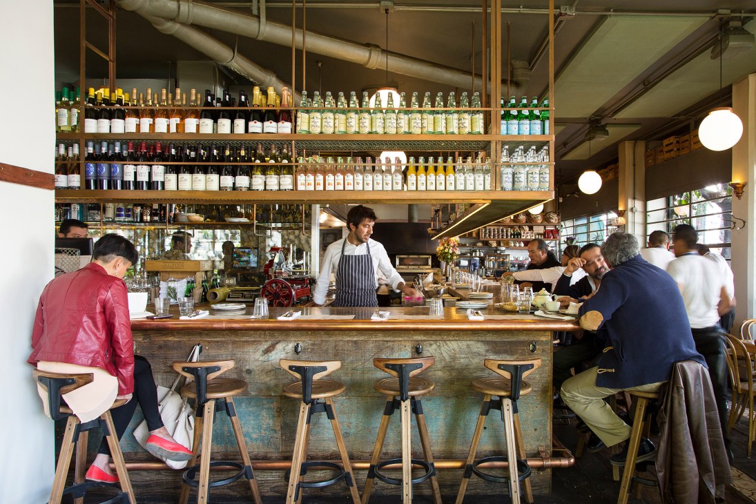 Interior of Lardo restaurant with bartender and diners at wooden counter beneath shelves of bottles.