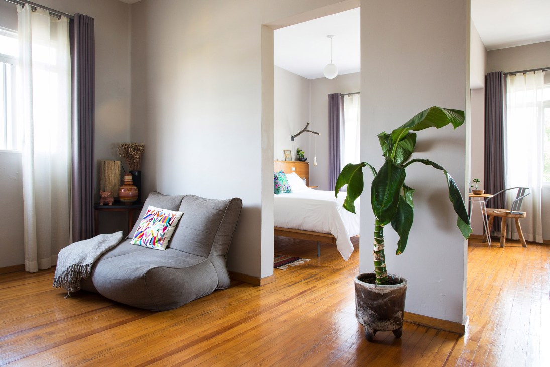 Modern interior space with grey curved chair, potted plant, and view into bedroom with white bedding.