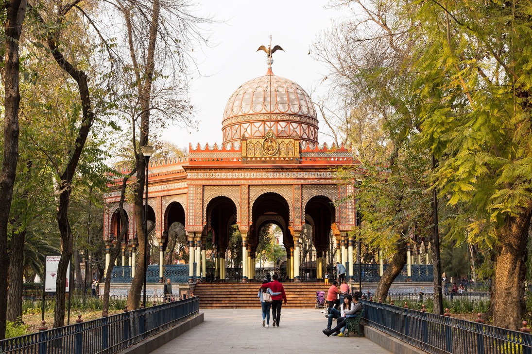 Orange and red domed structure with arches in a tree-lined public plaza with visitors walking.