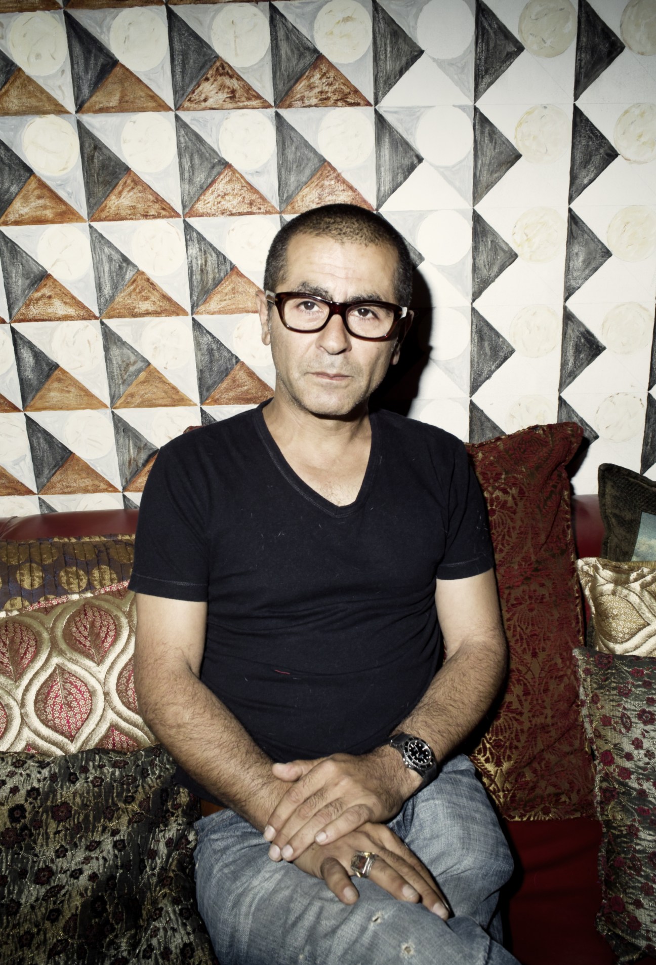 Man wearing glasses and black shirt sitting on patterned cushions against geometric wall.