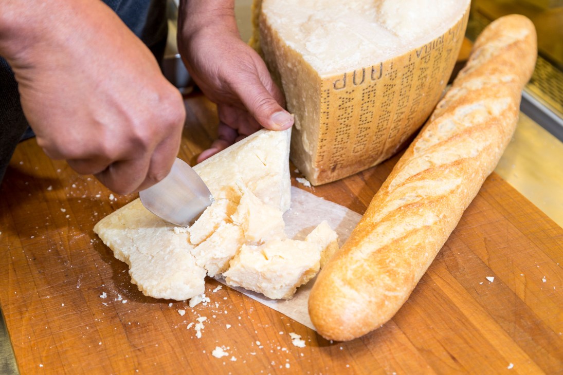 Hand cutting a wedge of Parmigiano Reggiano cheese on a wooden board with bread.