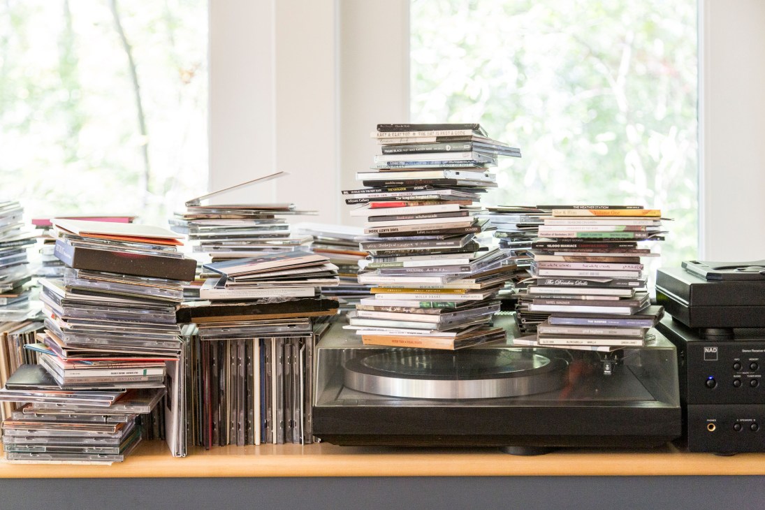 Stacks of CDs and music albums arranged on a wooden shelf next to a turntable and speaker.