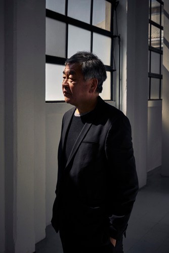 Architect Kengo Kuma stands in a minimalist white corridor with large windows.