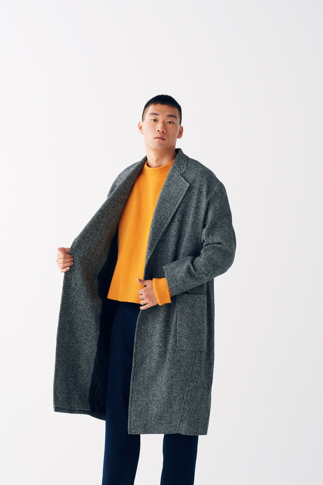 Man wearing charcoal herringbone overcoat with yellow cashmere knit and dark trousers.