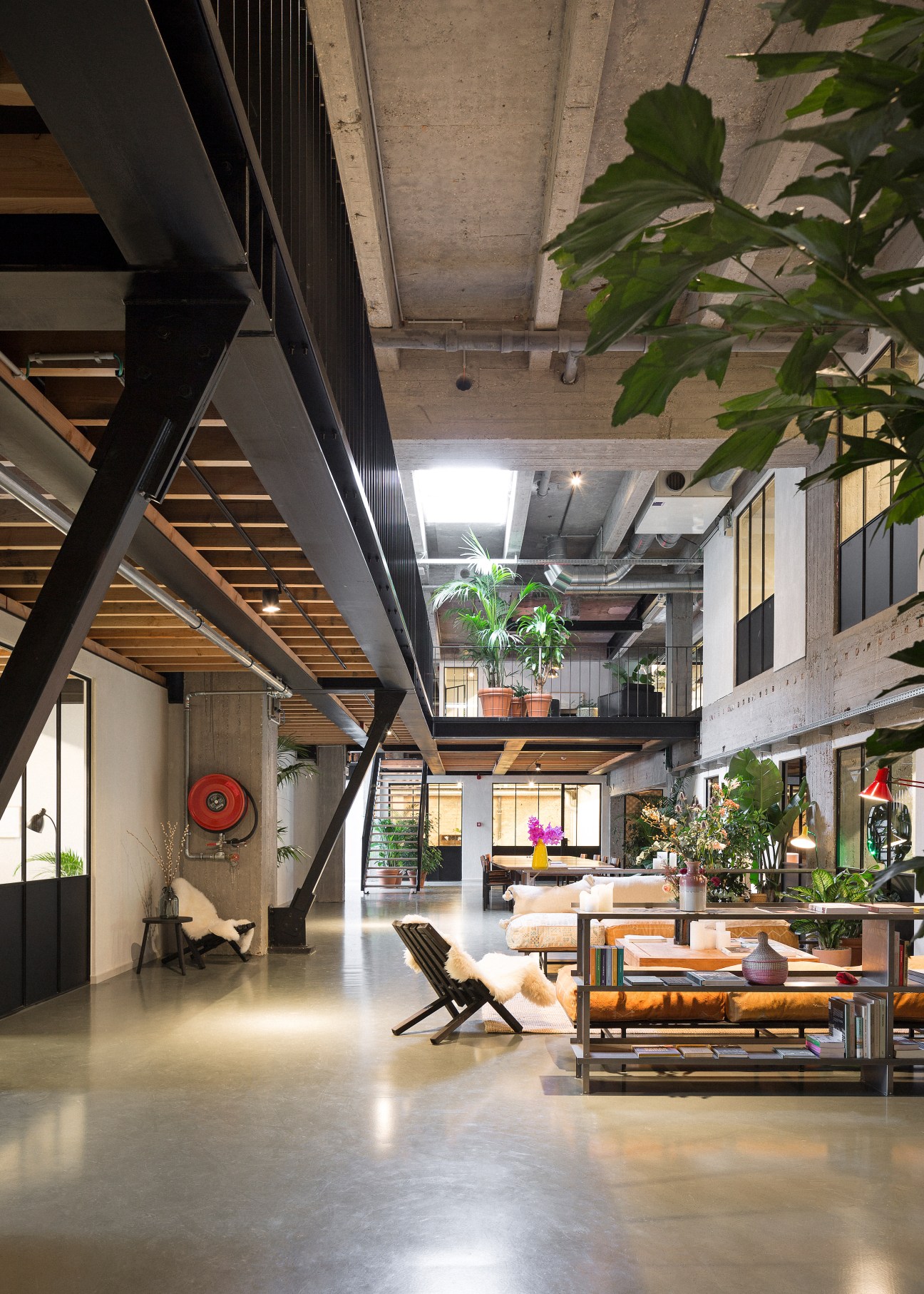 Modern open-plan office space with exposed concrete ceiling, wooden and steel structural elements, green plants, and contemporary furniture including seating areas.