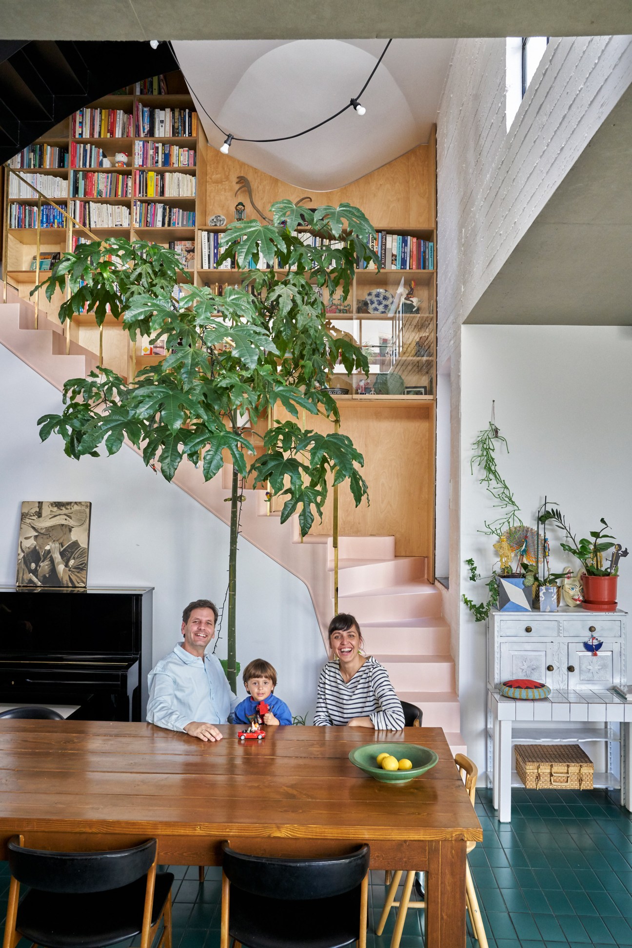 Family of three seated at wooden dining table in bright interior with pink staircase, bookshelves, and potted plant behind them.