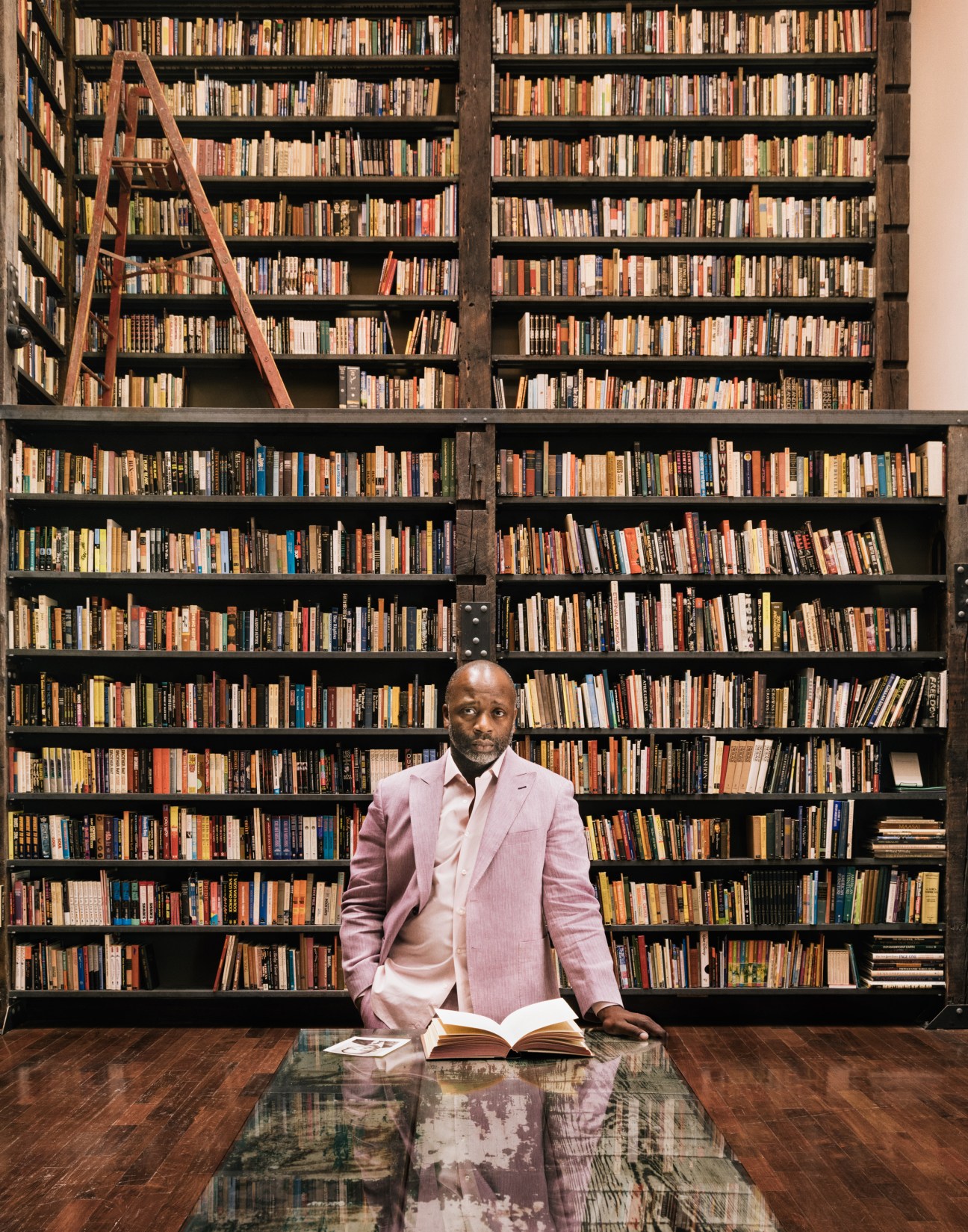 Theaster Gates stands at a table with an open book in front of extensive floor-to-ceiling bookshelves at the Stony Island Arts Bank in Chicago.