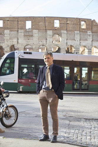 Paolo Arsena stands on a Roman street with a tram and ancient ruins in the background.