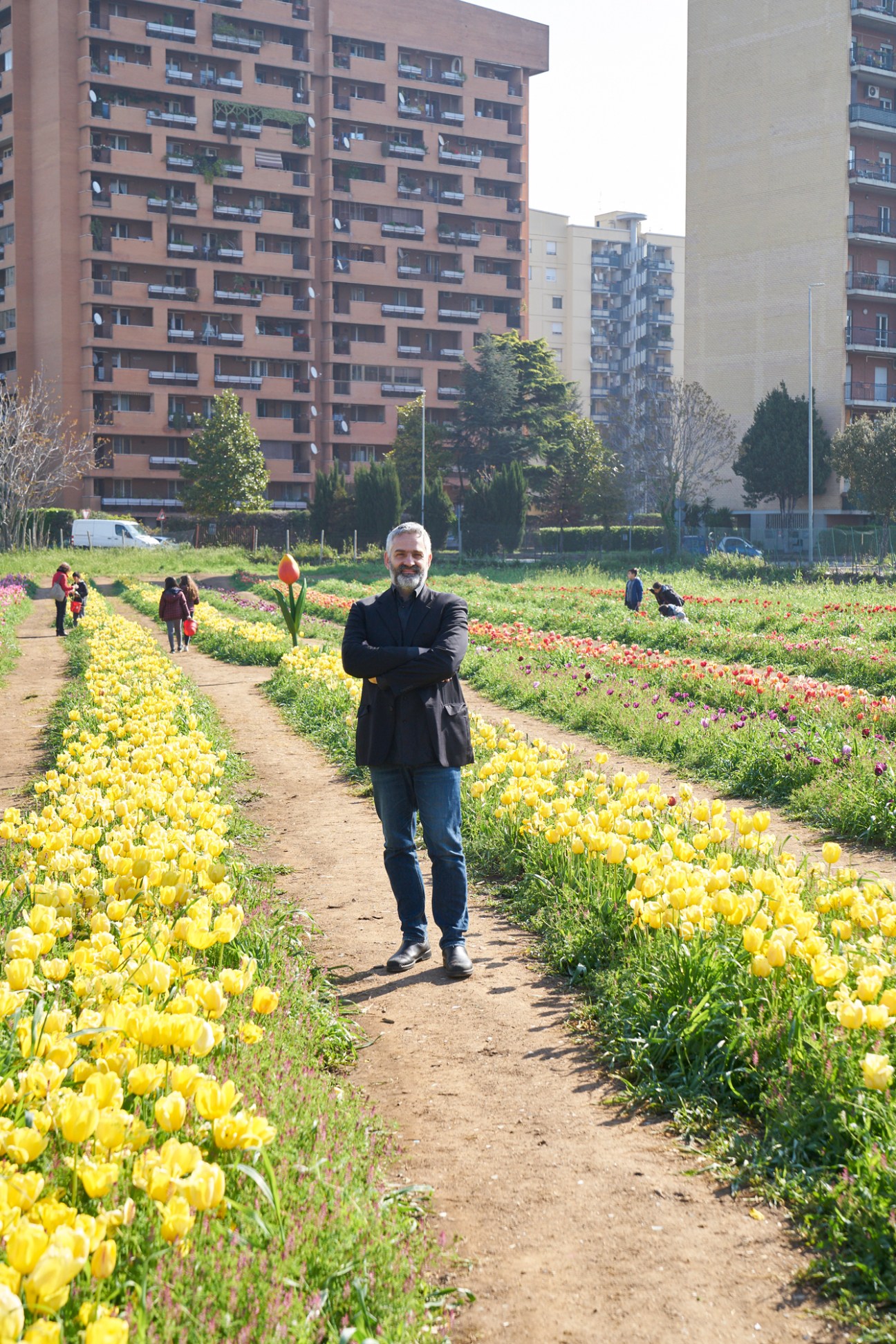 Claudio Gnessi stands in a flower garden with yellow and purple tulips, surrounded by apartment buildings in Rome's suburbs.