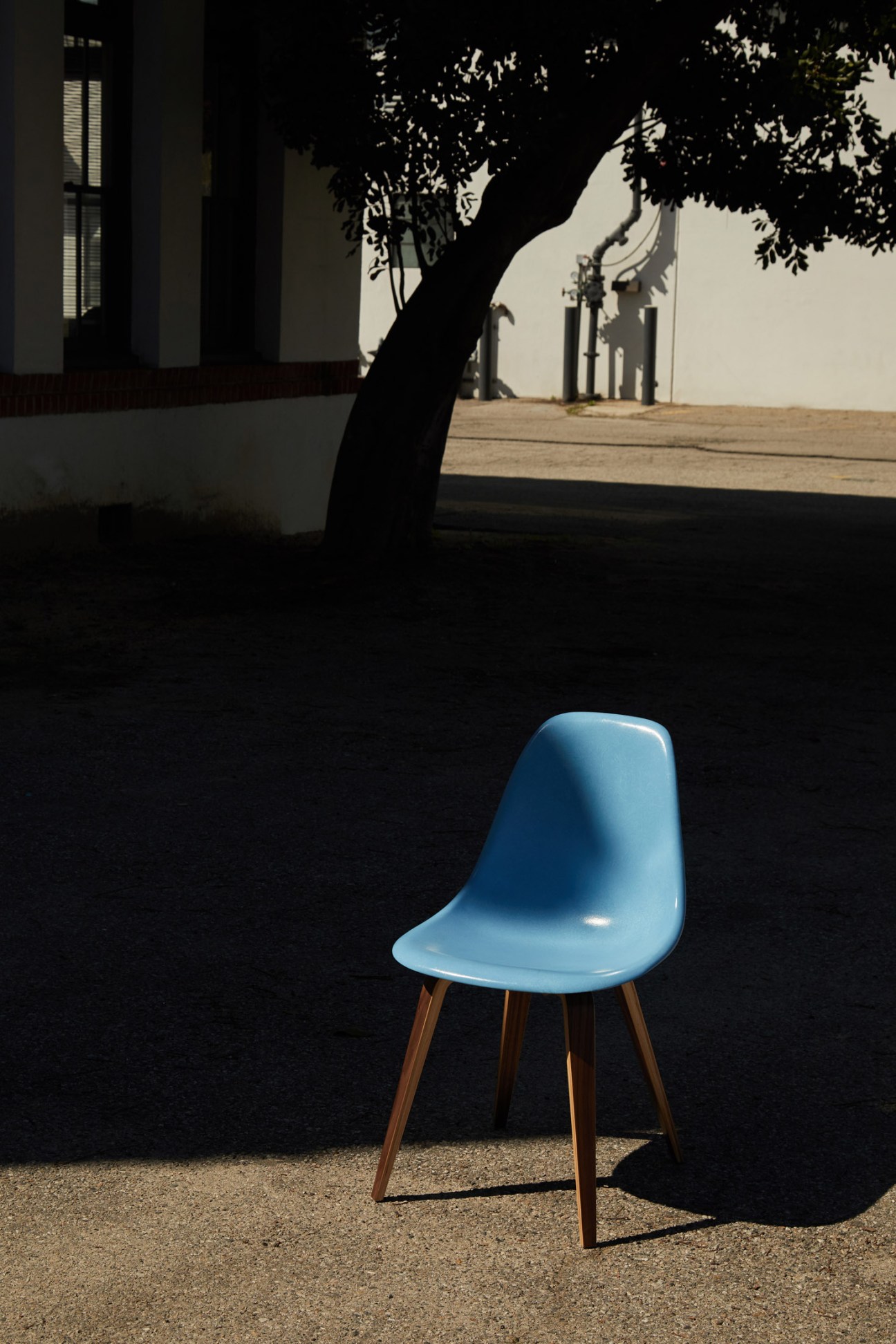 Light blue Eames Fiberglass chair with wooden legs in a courtyard with shadow of tree and white building.